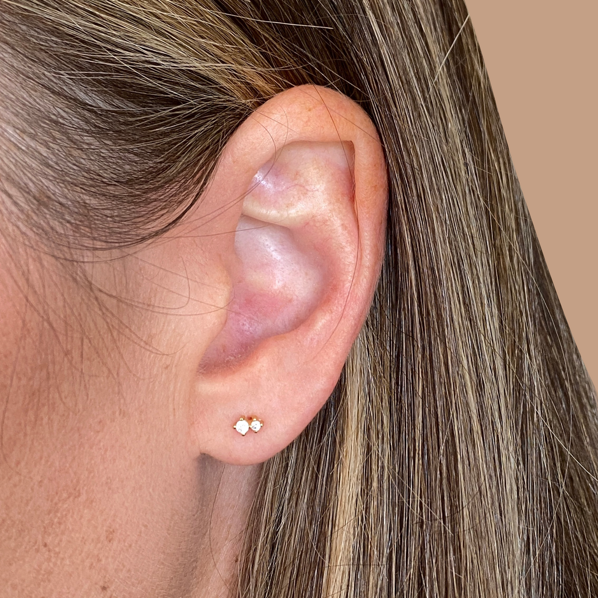 Close-up of an ear with a small crystal stud gold earring against a neutral background