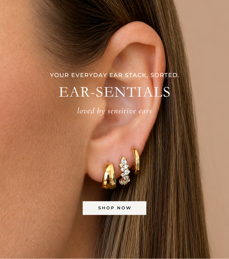 Close-up of a woman's ear wearing three gold earrings, including a small cluster with clear stones, on a warm beige background with overlaid text reading 'EAR-SENTIALS' and a 'SHOP NOW' button.