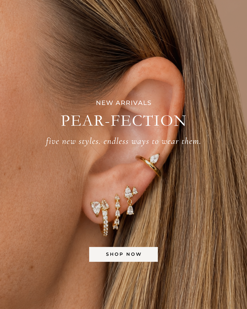 Close-up of a woman's ear wearing multiple gold and clear crystal ear piercings, with overlaid text reading 'NEW ARRIVALS PEAR-FECTION five new styles. endless ways to wear them.